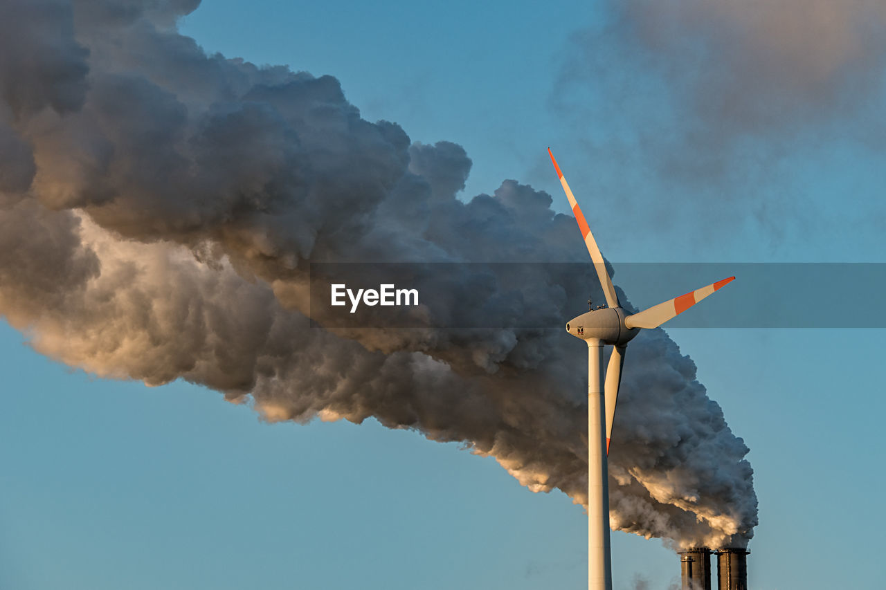 Low angle view of wind turbine by smoke emitting from chimney against blue sky