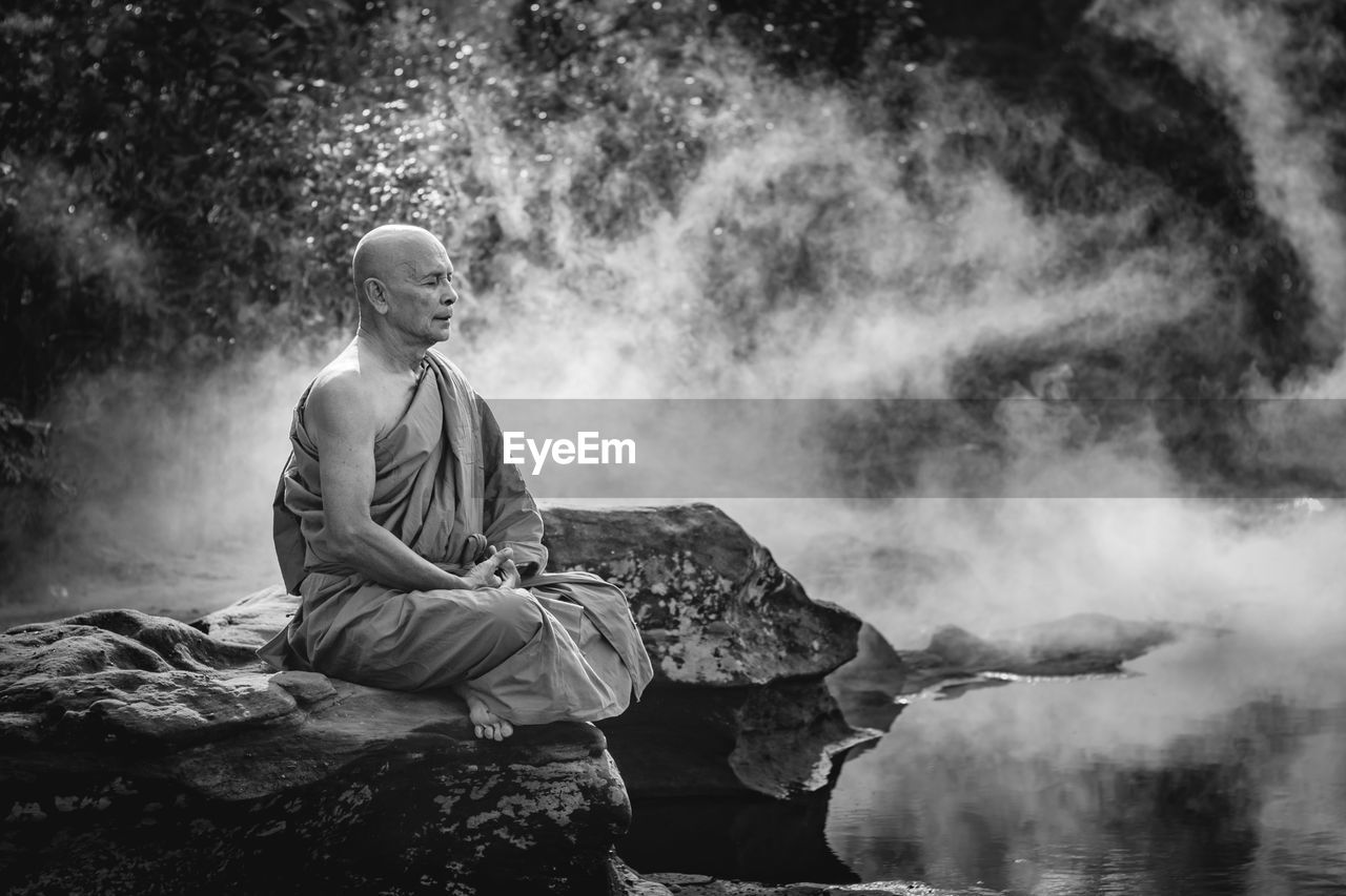 Full length of monk meditating while sitting on rock by river