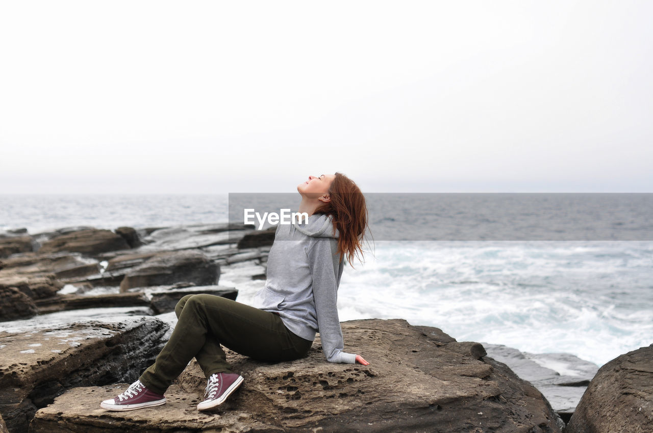 Young woman in hoodie, jeans sits on rocks near the stormy oceans with waves. local tourism