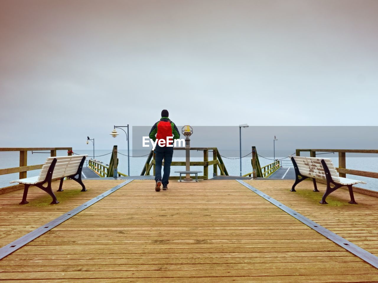 Tourist with red backpack on wooden pier above sea. man in trekking suit in harbor in rainy day.