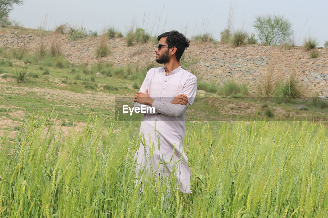 plant, one person, agriculture, adult, field, nature, land, grass, landscape, crop, growth, rural scene, grassland, men, young adult, prairie, meadow, environment, casual clothing, standing, natural environment, three quarter length, rural area, occupation, day, sky, looking, beauty in nature, outdoors, tranquility, leisure activity, lifestyles, paddy field, cereal plant, green, scenics - nature, front view, holding, clothing, tranquil scene, smiling, non-urban scene