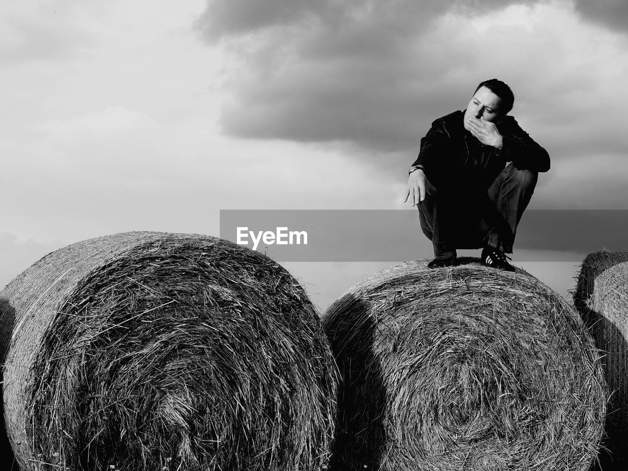 Full length of man crouching on hay bale against cloudy sky