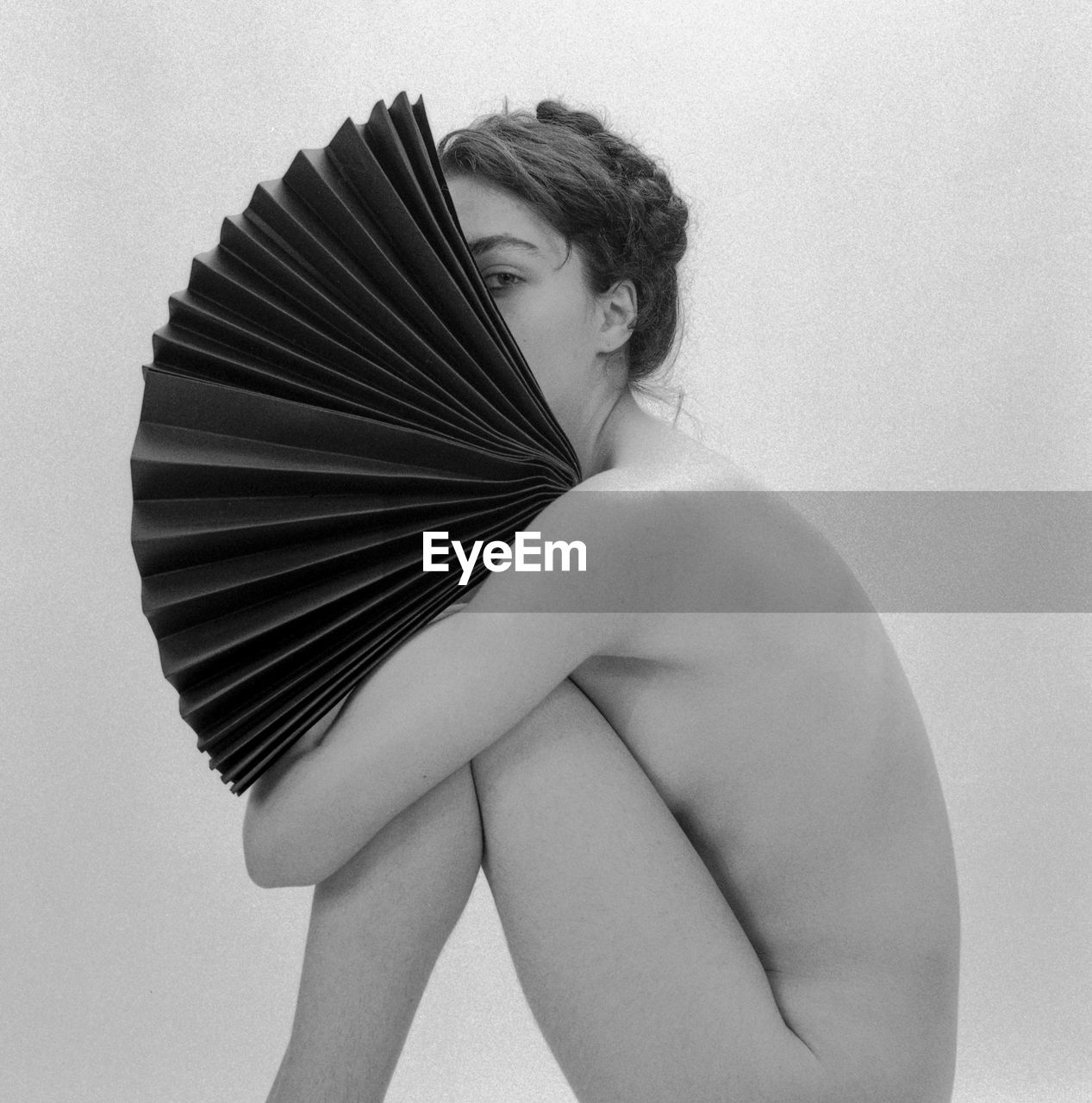 Side view of naked woman holding hand fan sitting against white background