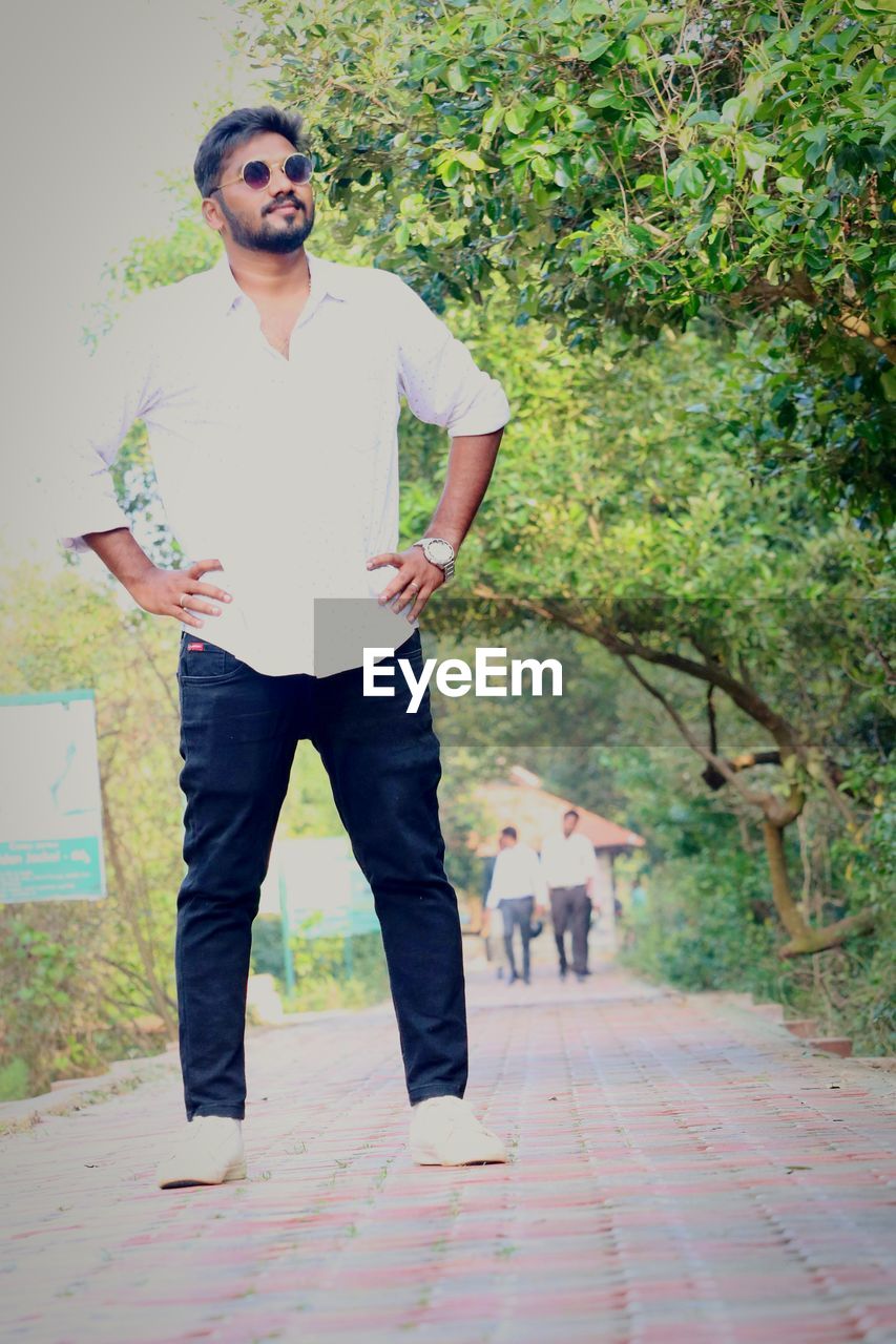 one person, full length, adult, spring, front view, young adult, standing, men, casual clothing, portrait, fashion, plant, day, glasses, green, footpath, lifestyles, sunglasses, nature, trousers, tree, leisure activity, outdoors, emotion, looking at camera, photo shoot, beard, looking, facial hair, smiling, happiness