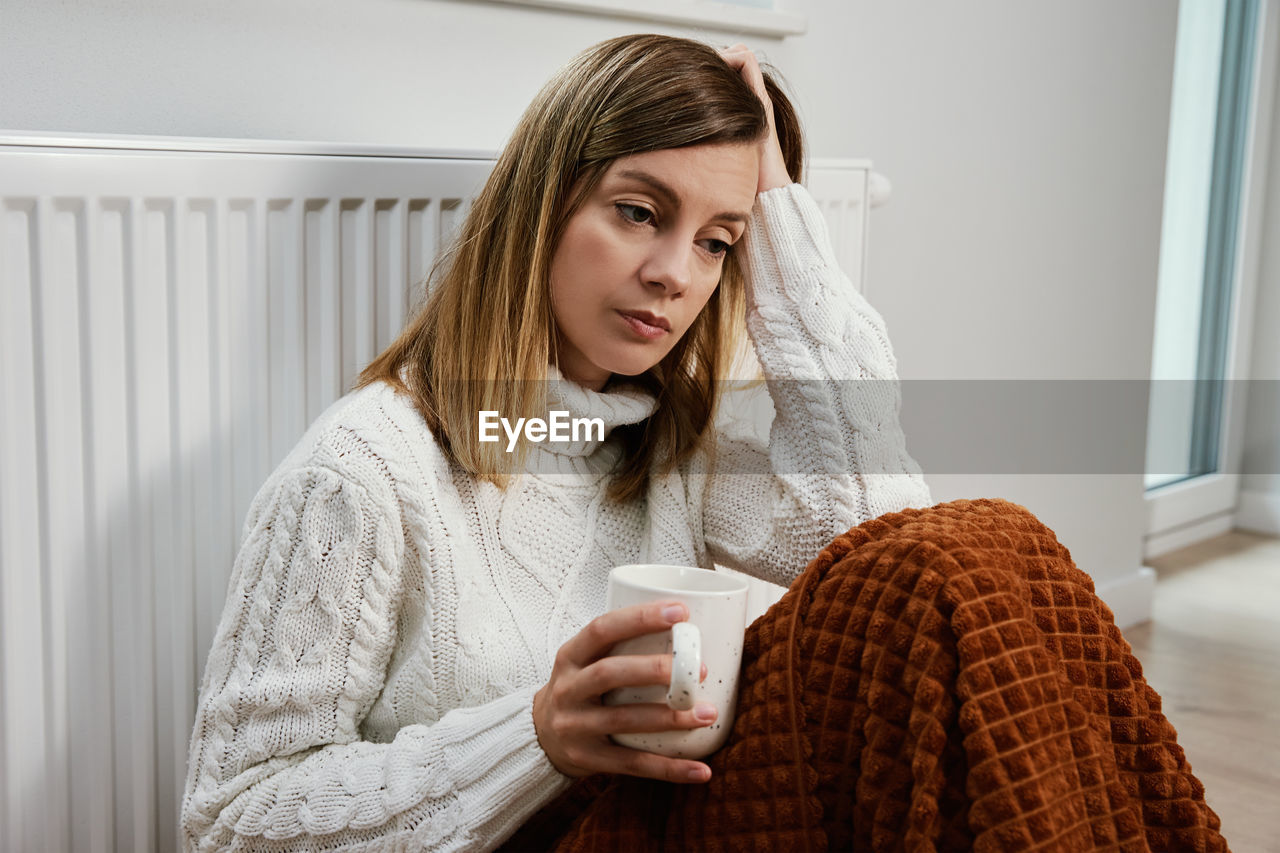 Worried woman sit near heating radiator under blanket with cup of tea
