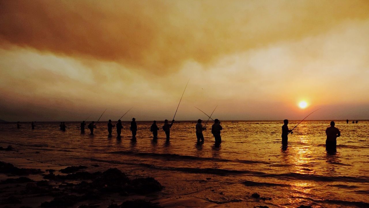 Scenic view of people fishing in sea against cloudy sky at sunset