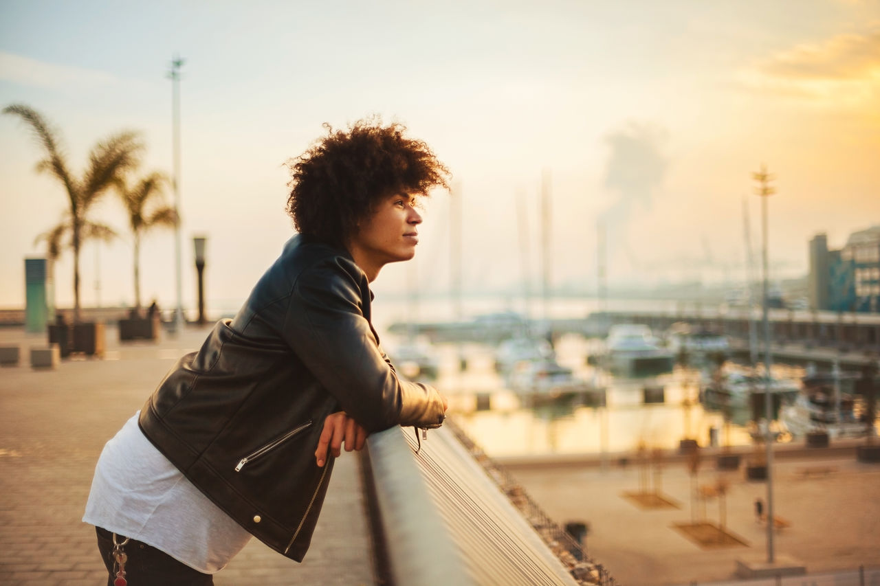 Young man looking away standing by railing during sunset