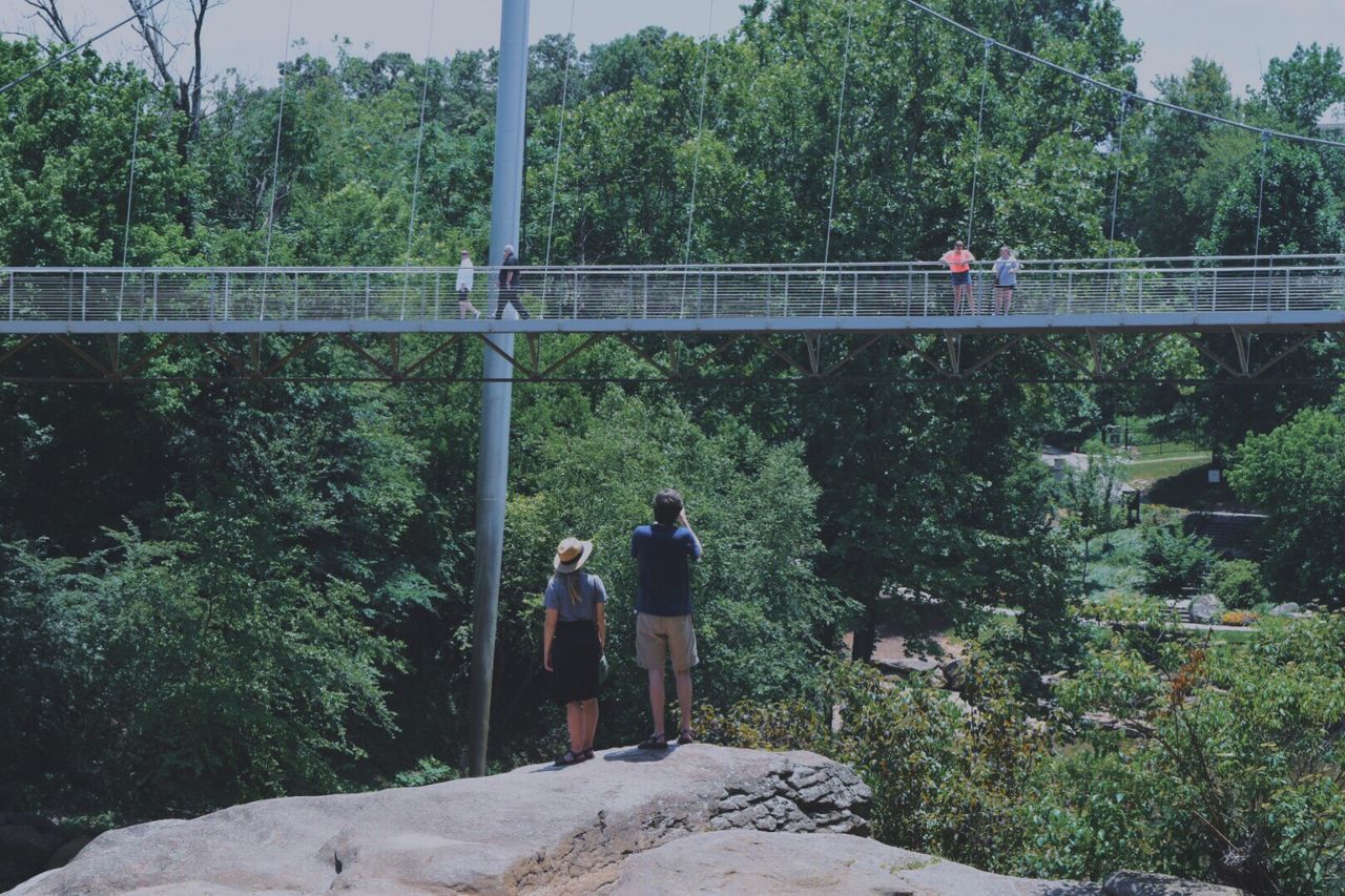 Rear view full length of friends standing on rock against footbridge