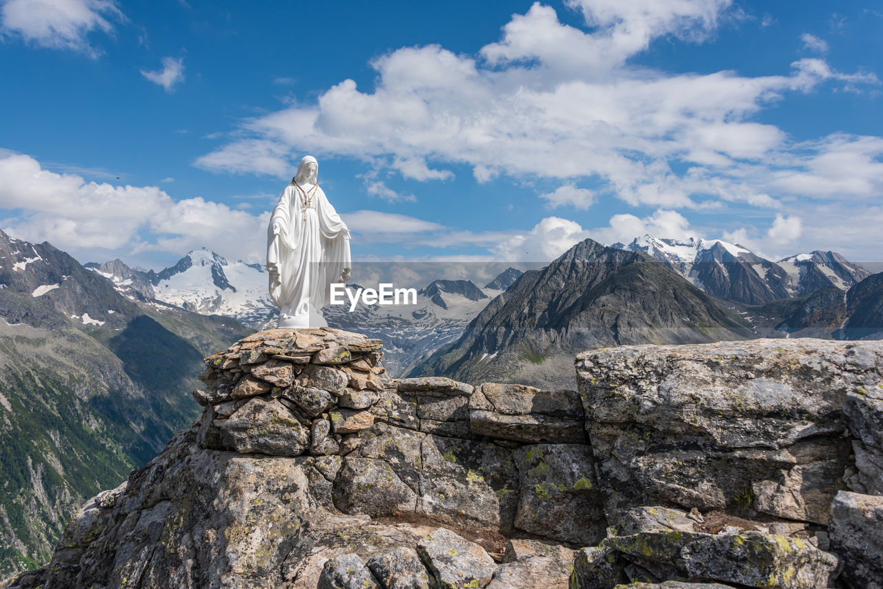White statue of virgin mary, mother of god, placed on top of the mountains, blue sky, white clouds.