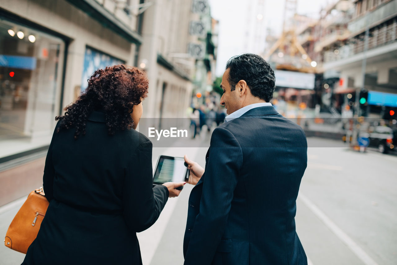 Rear view of businesswoman showing digital tablet to businessman while walking on sidewalk in city