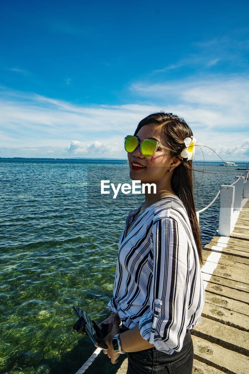 Young woman wearing sunglasses standing by sea against sky