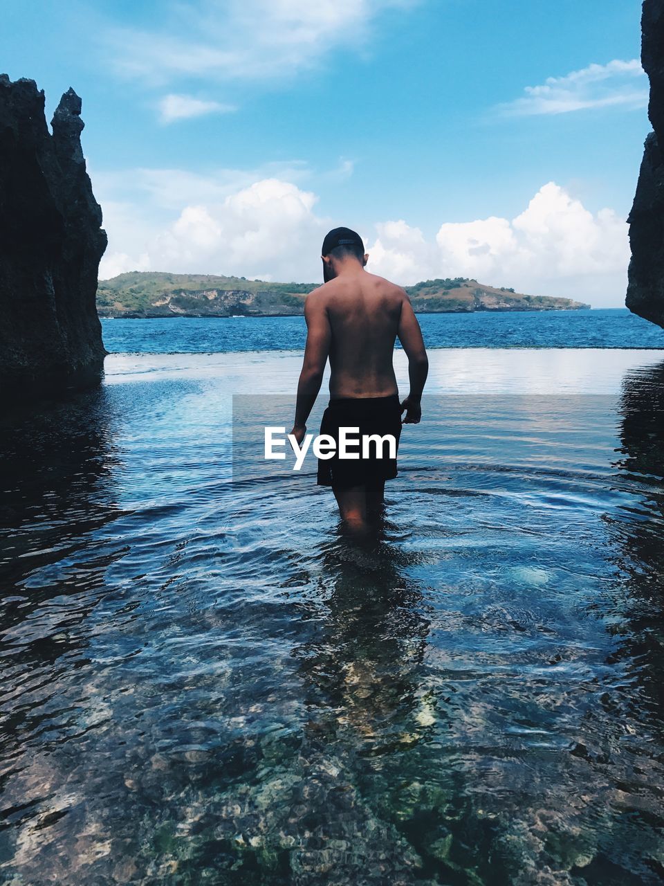 REAR VIEW OF SHIRTLESS MAN STANDING ON ROCK IN SEA