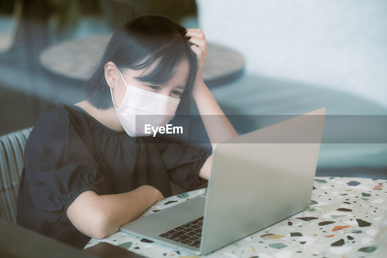 Woman with head in hands wearing while mask sitting by laptop on table