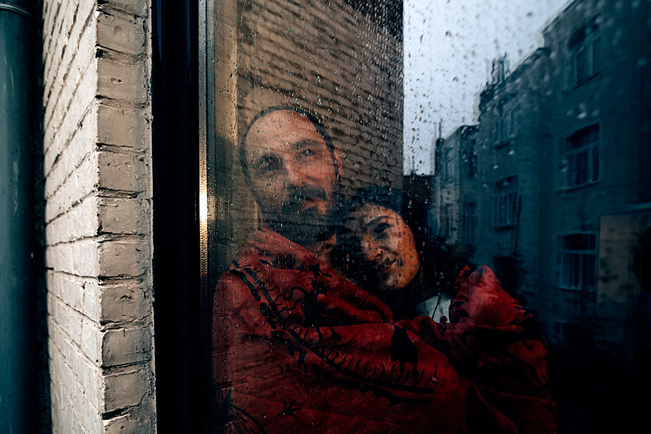 Couple wrapped in a red blanket looking outside from a backyard door window on a grey rainy day
