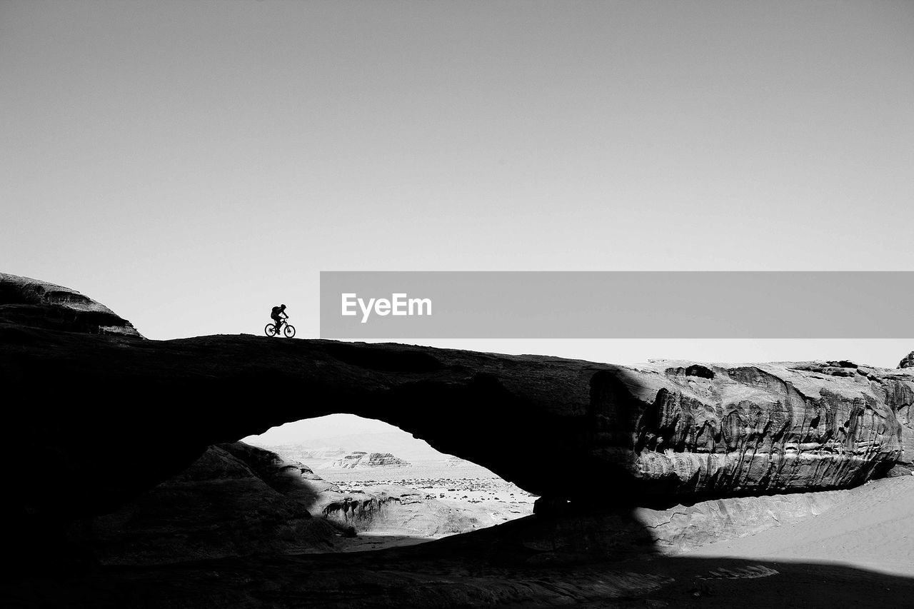 Side view of silhouette person bicycling on rocky land against clear sky
