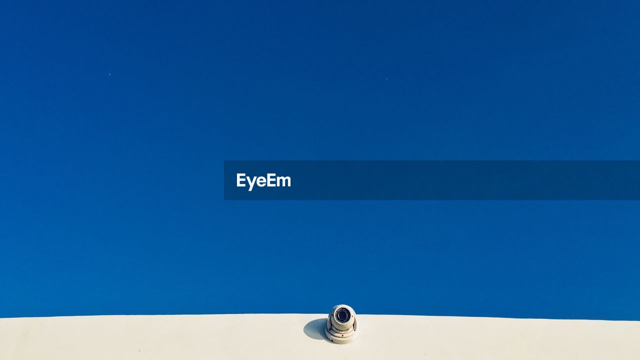 Low angle view of security camera on wall against blue sky