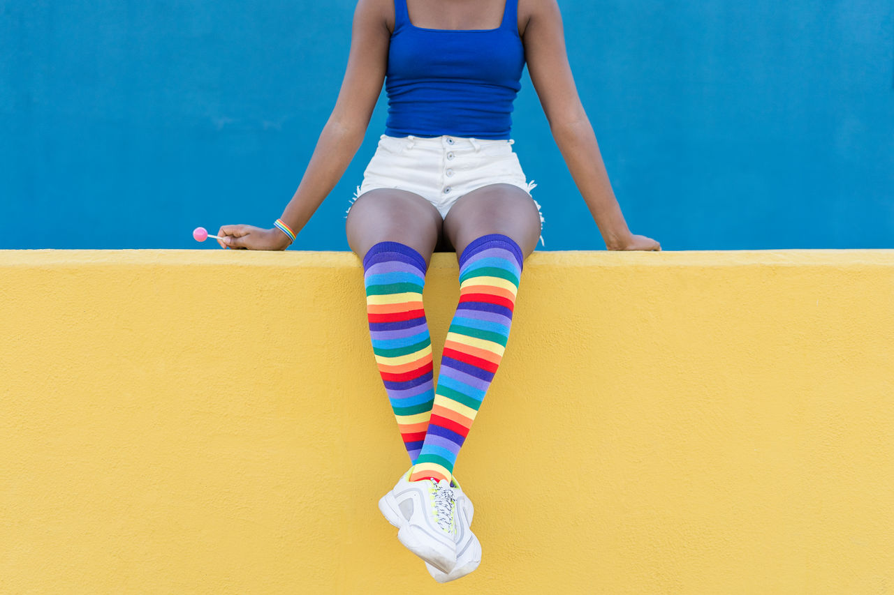 Crop view of anonymous black female in colorful clothes sitting on yellow barrier against blue wall on street while holding a lollypop