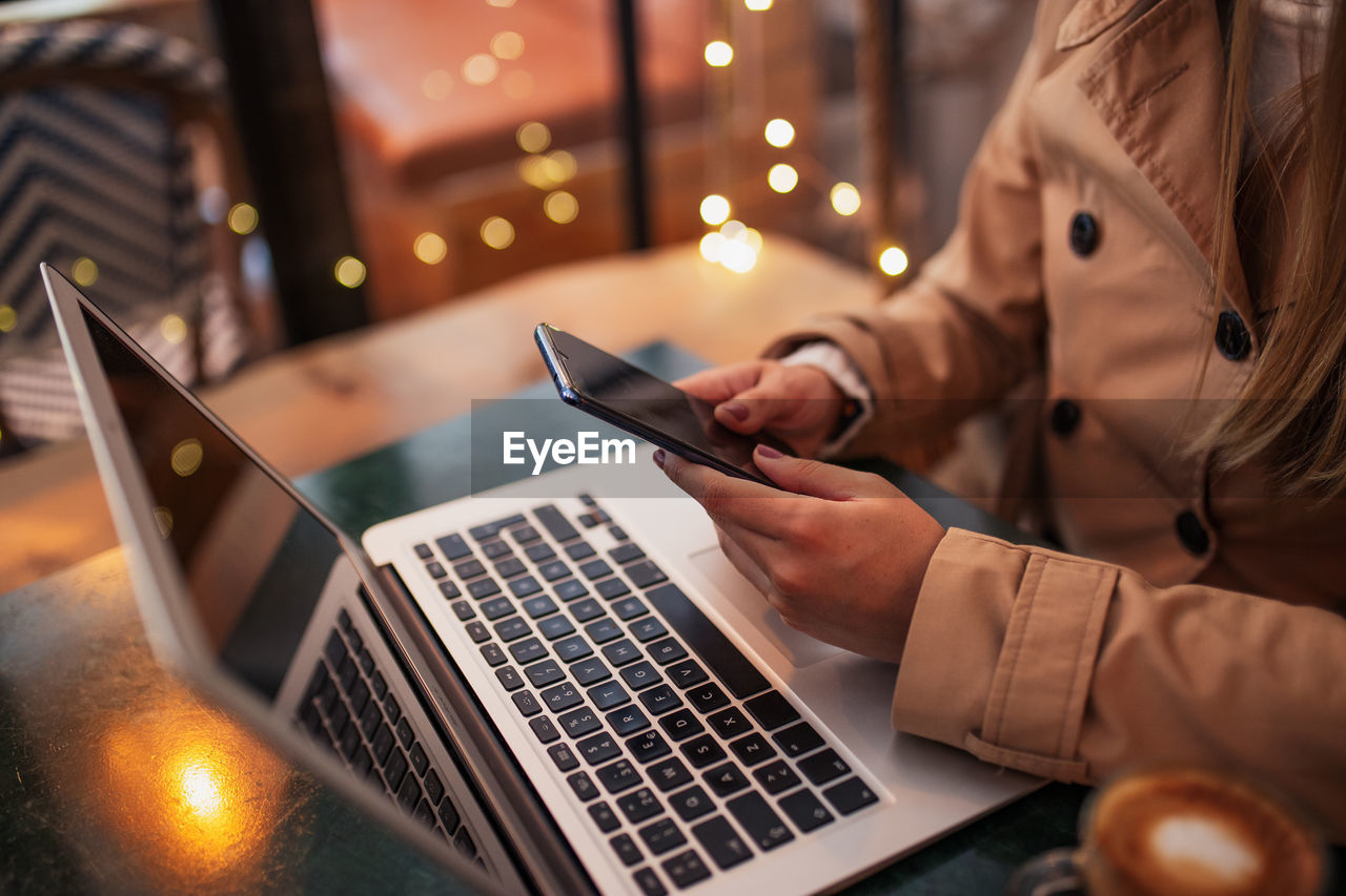 High angle of cropped unrecognizable young female freelancer messaging on mobile phone while sitting at table with laptop and cup of coffee in cozy cafe and working or remote project