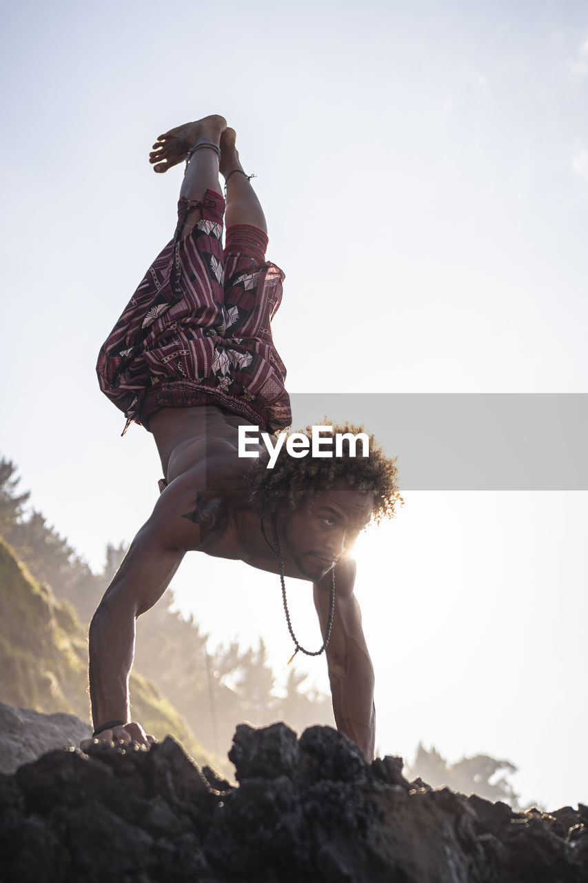 Young man looking away while practicing handstand on rock at beach against sky