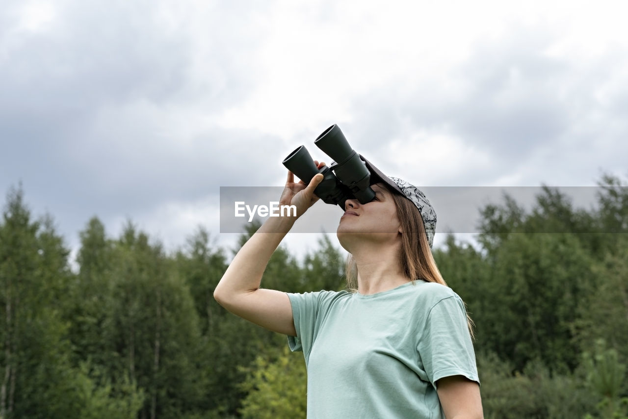 Young blonde woman bird watcher in cap  looking through binoculars cloudy sky in forest ornithology