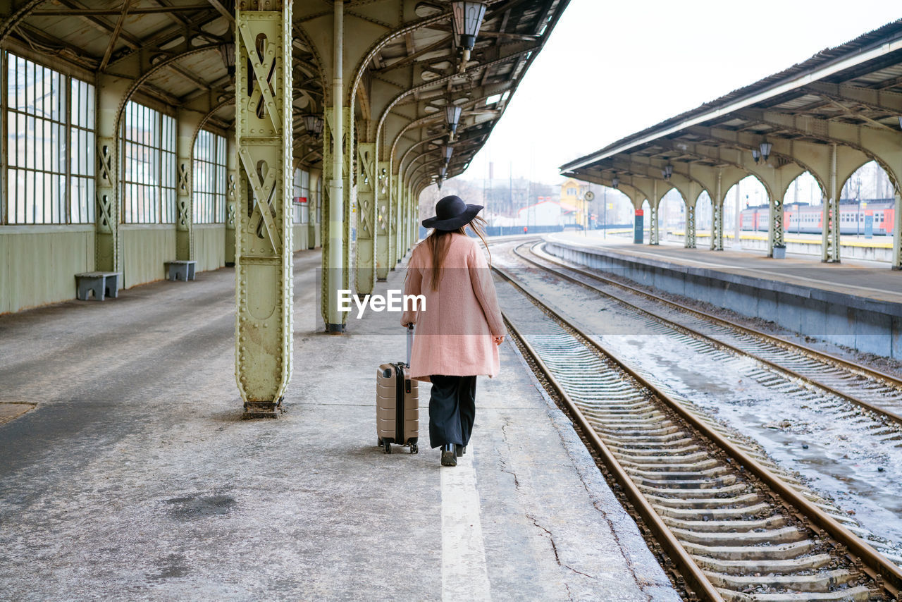 Woman in pink coat walks with suitcase on an empty platform with her back