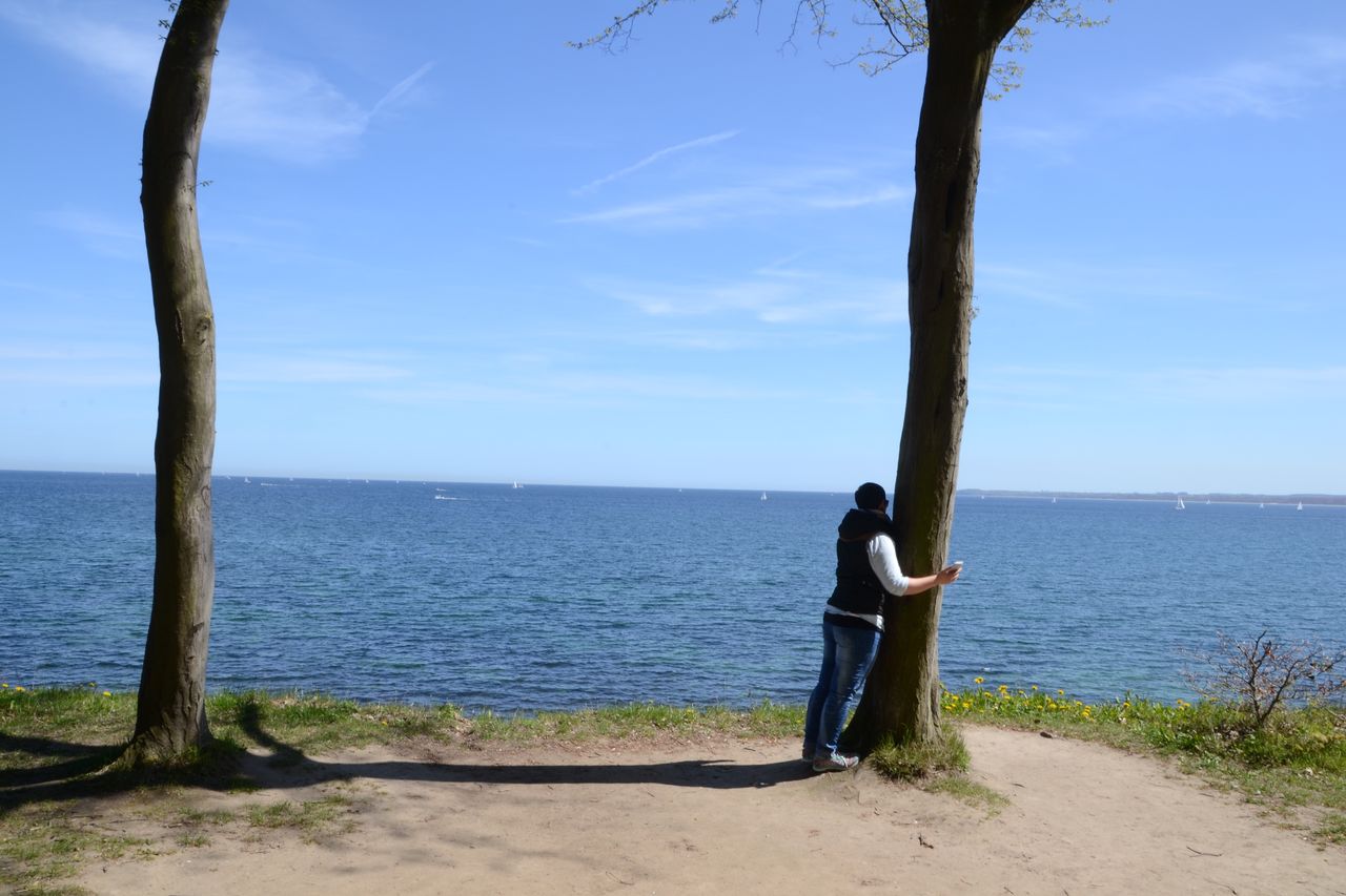 Full length of man embracing tree trunk while looking at sea against blue sky