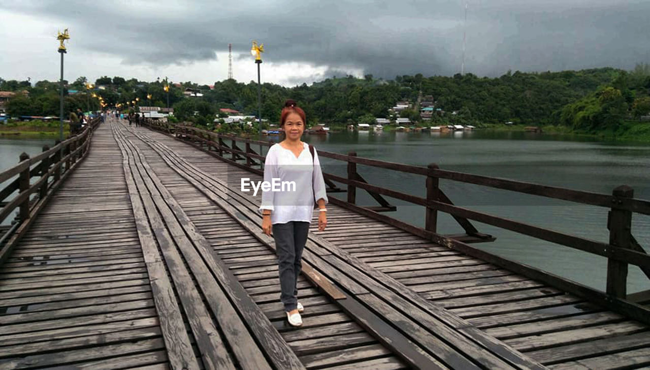 Portrait of woman on wooden footbridge over lake against cloudy sky