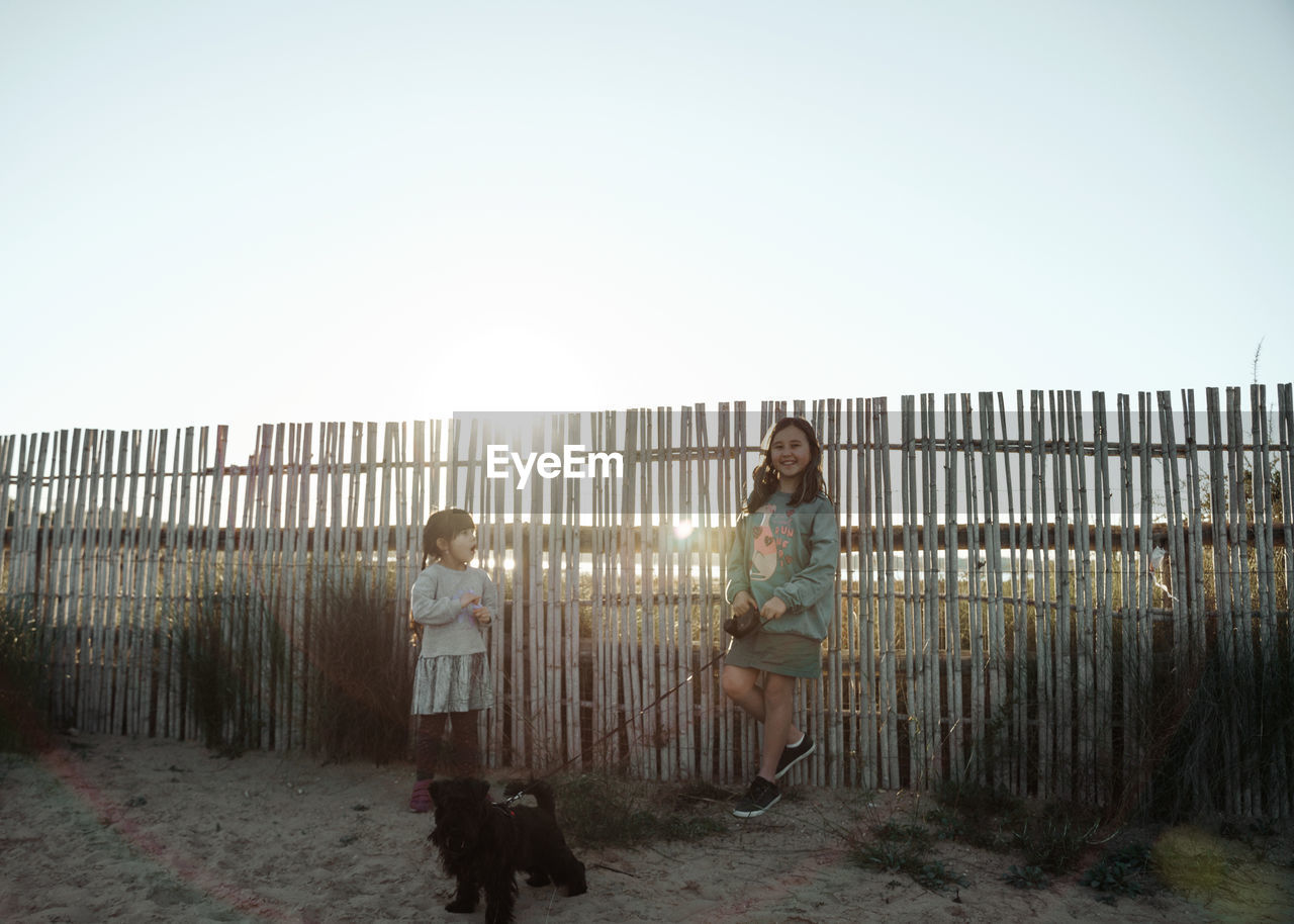 Rear view of two girl standing by fence with dog against clear sky