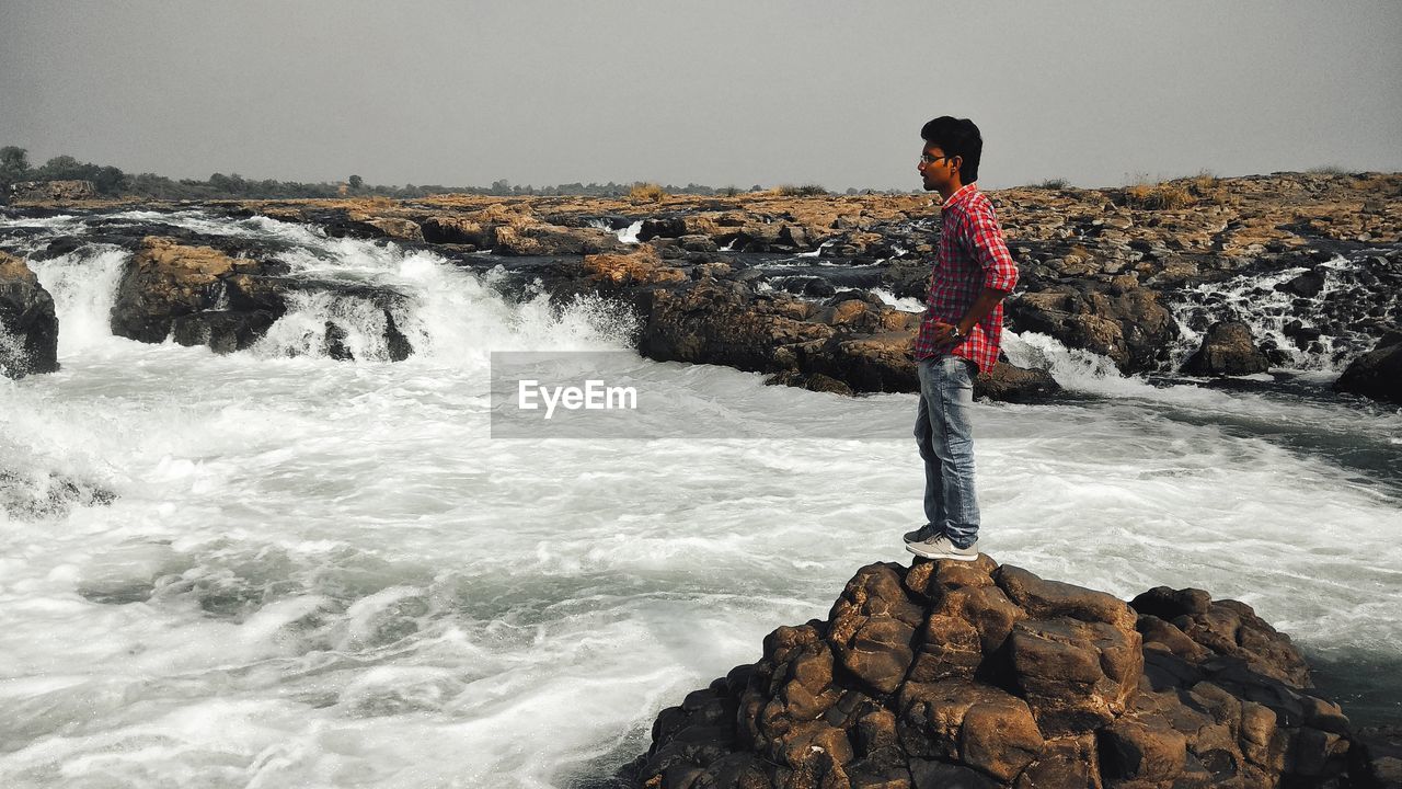 Side view of man standing on rock by sea against clear sky