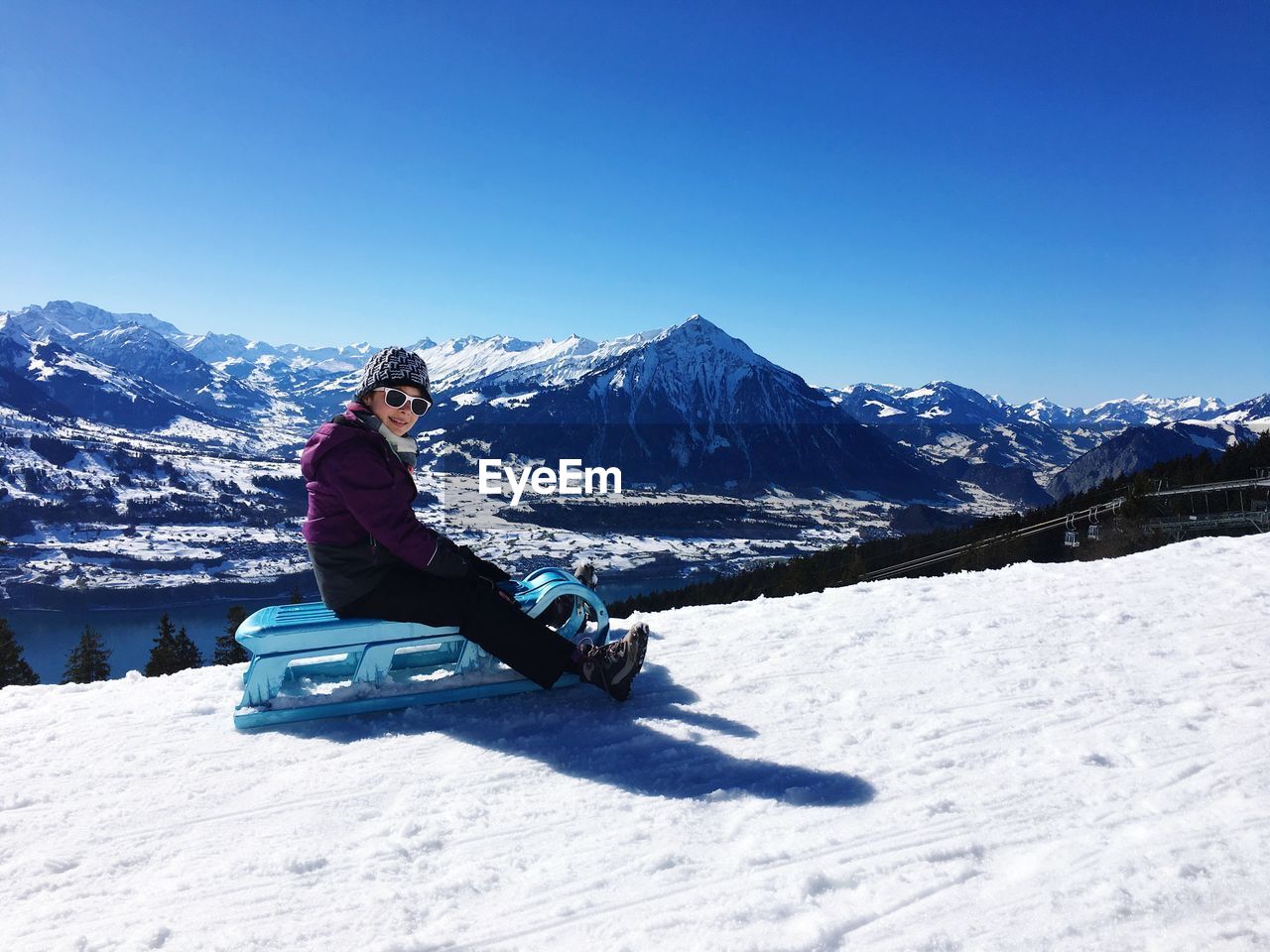 Woman sitting over sled on snow covered land against mountains and clear blue sky