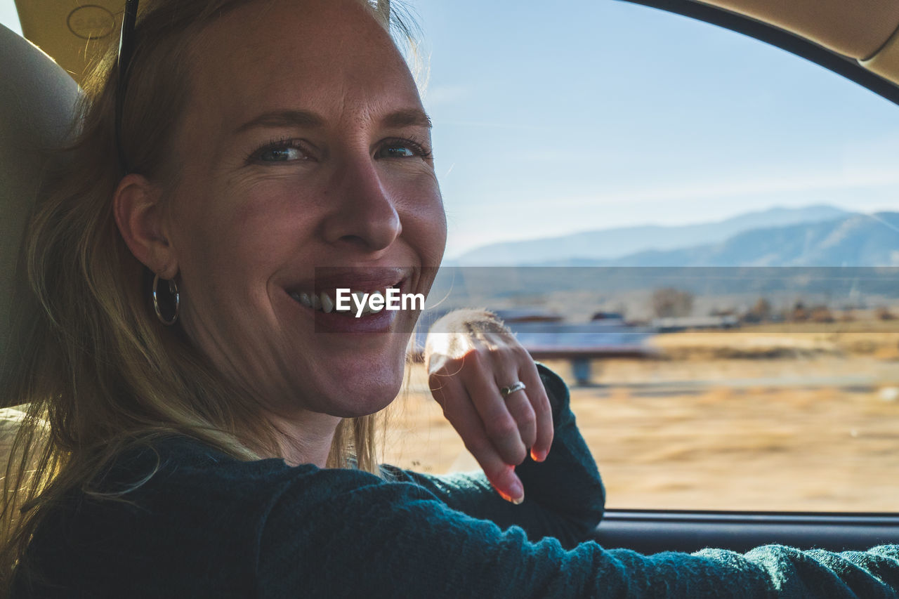 Smiling woman looks at camera while driving on road trip, view of desert out of drivers window