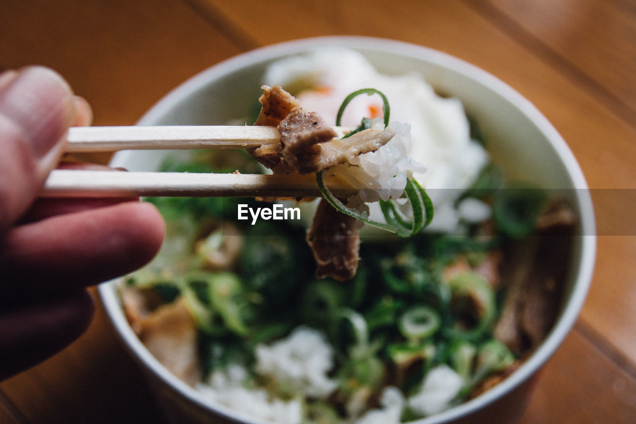 Cropped hand of person holding food in chopsticks against bowl
