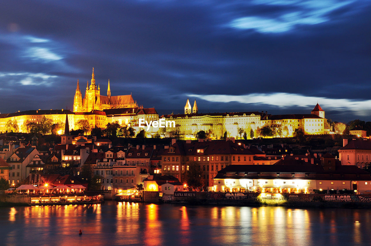 River in front of illuminated town against cloudy sky at dusk