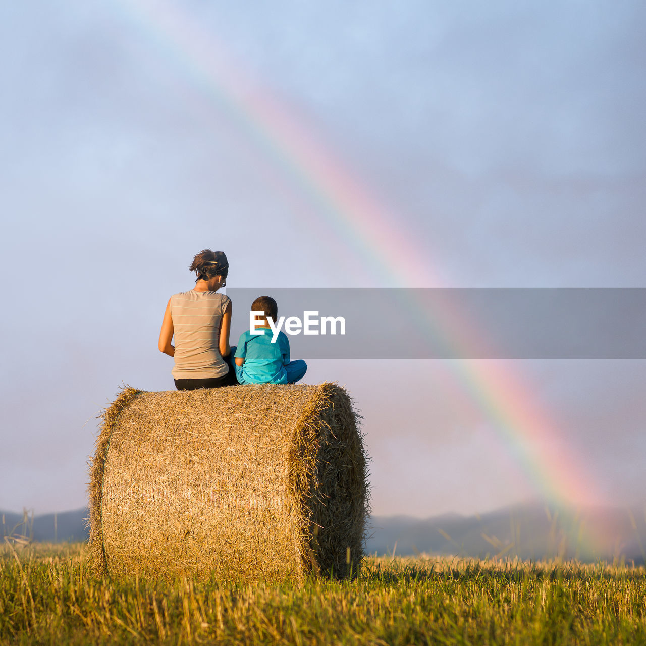 Rear view of mother and son sitting on hay bale against sky during sunset
