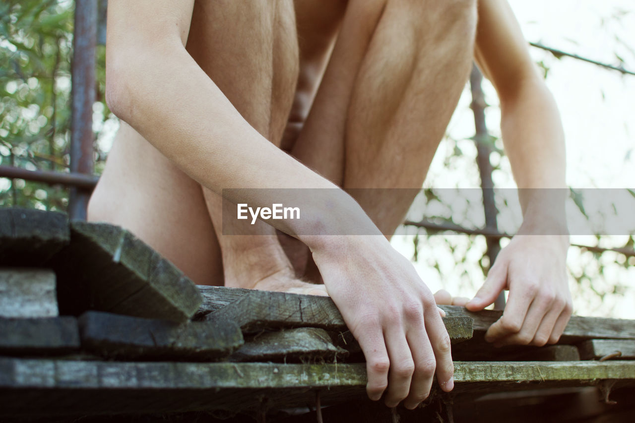 Low section of naked man sitting on wooden plank outdoors