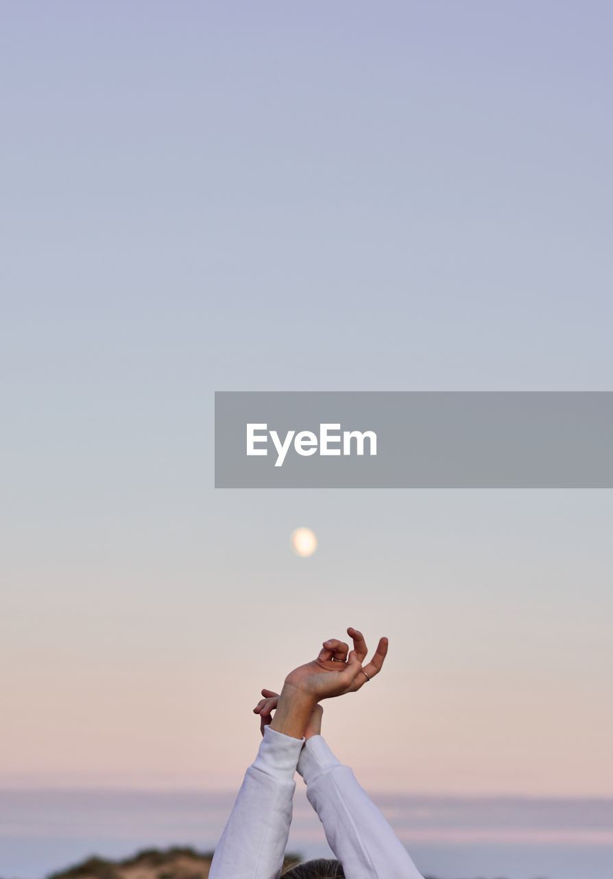 Unrecognizable crop female standing with raised arms on background of sunset sky with moon and enjoying freedom during vacation in summer