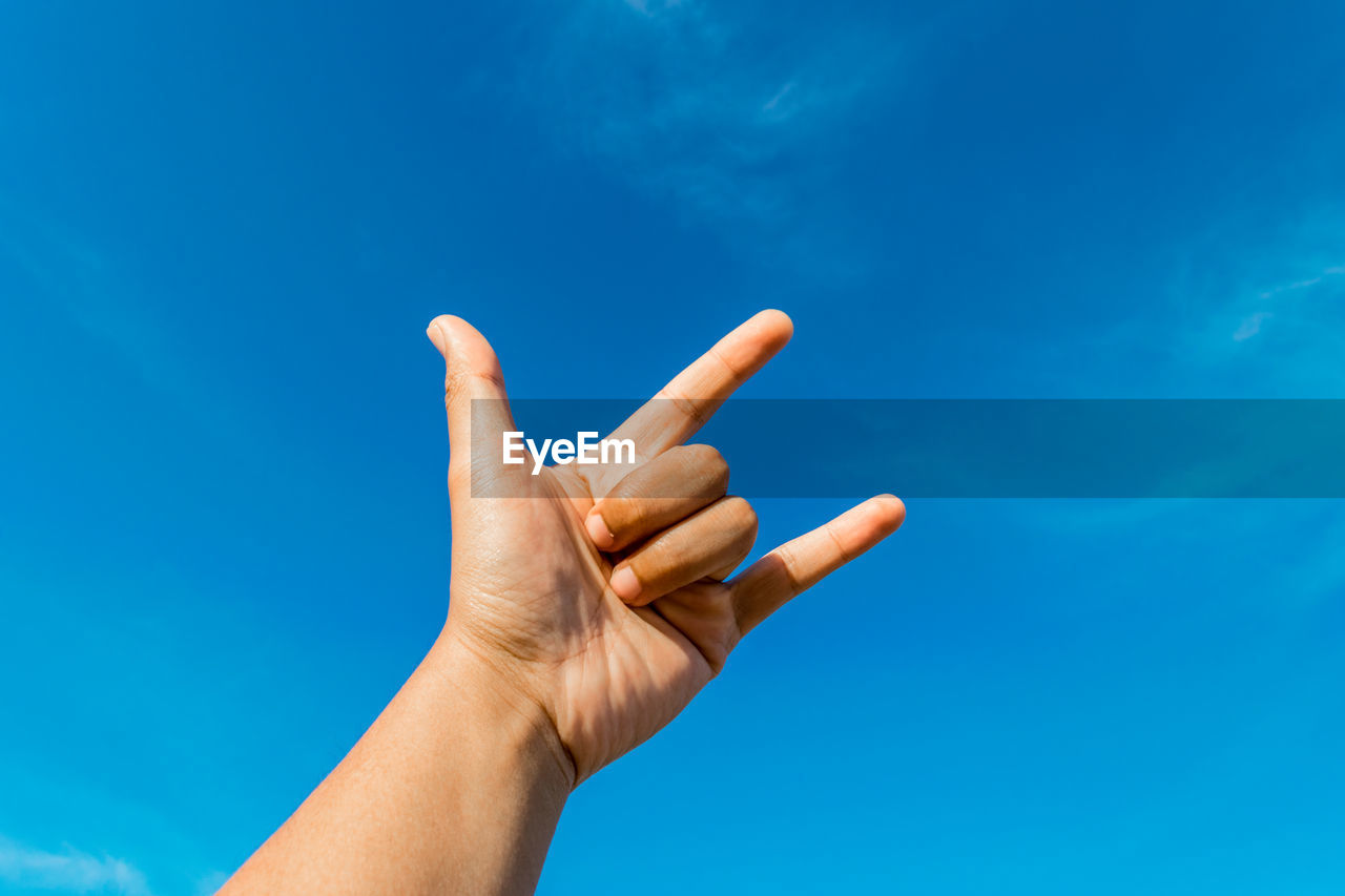 Cropped hand gesturing horn sign against blue sky