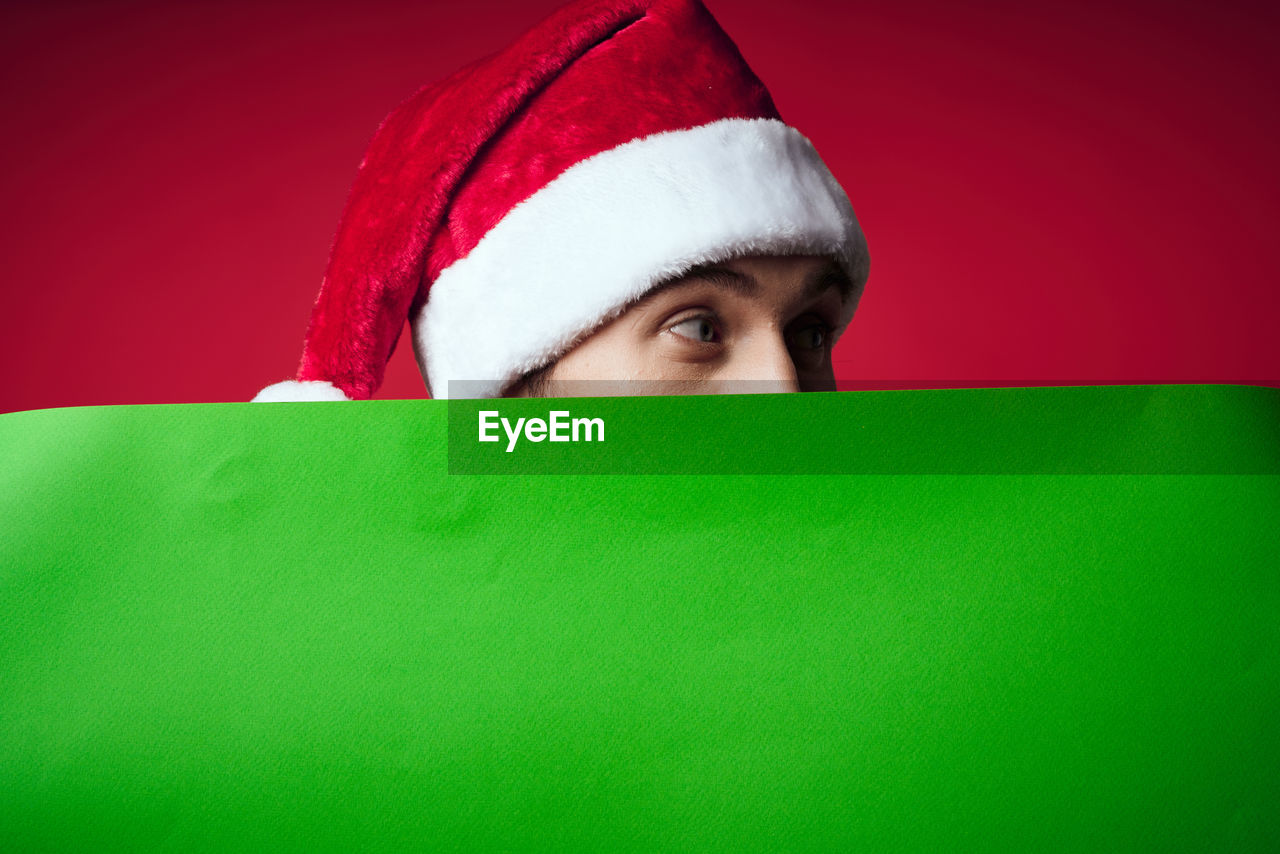 green, christmas, celebration, one person, portrait, surprise, holiday, santa hat, red, hat, studio shot, indoors, headshot, hiding, adult, human face, peeking, santa claus, looking at camera, emotion, young adult, clothing, colored background, copy space, gift, fun, costume, humor, paper, front view, looking, close-up