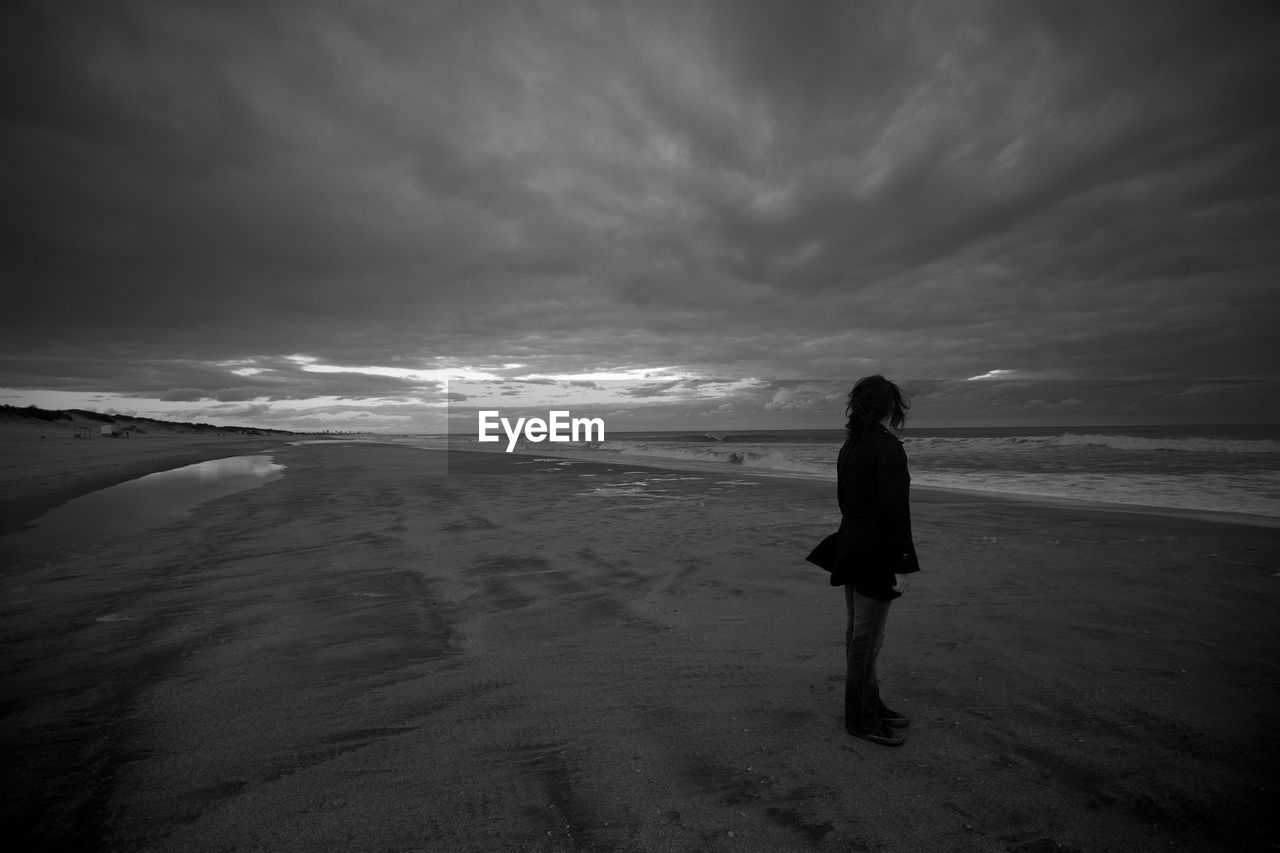 Woman standing at beach against cloudy sky during sunset