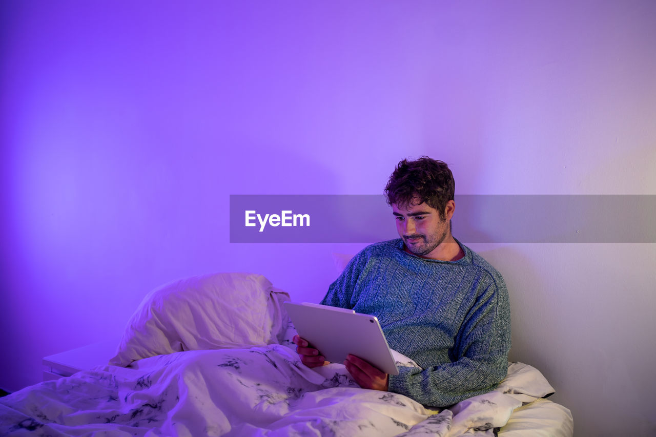 Man using digital tablet sitting on bed against wall at home