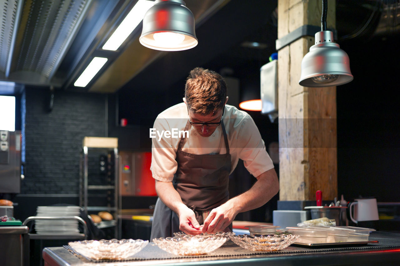 Young serious chef man in cooking apron serving meal in dishes on restaurant kitchen