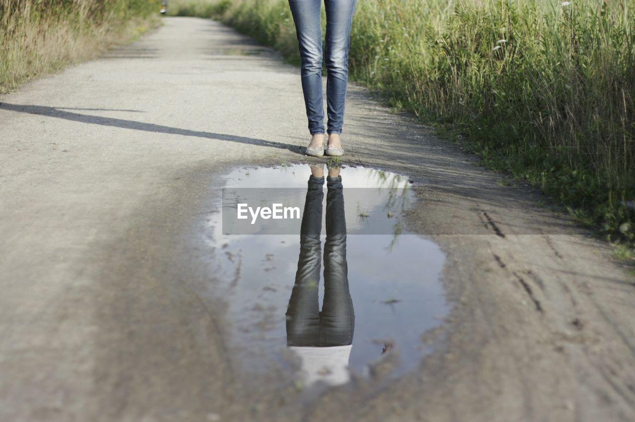 Low section of woman with reflection in puddle on footpath amidst meadow