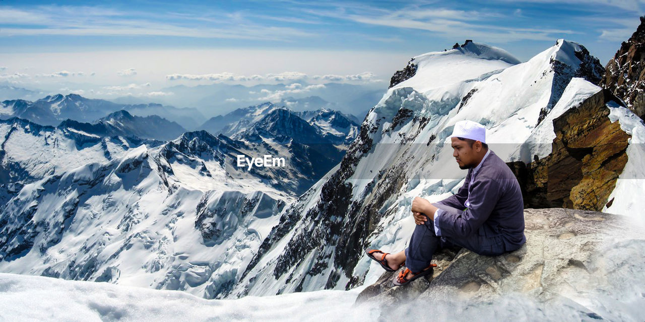 Side view of young man looking away while sitting on snowcapped mountain against sky