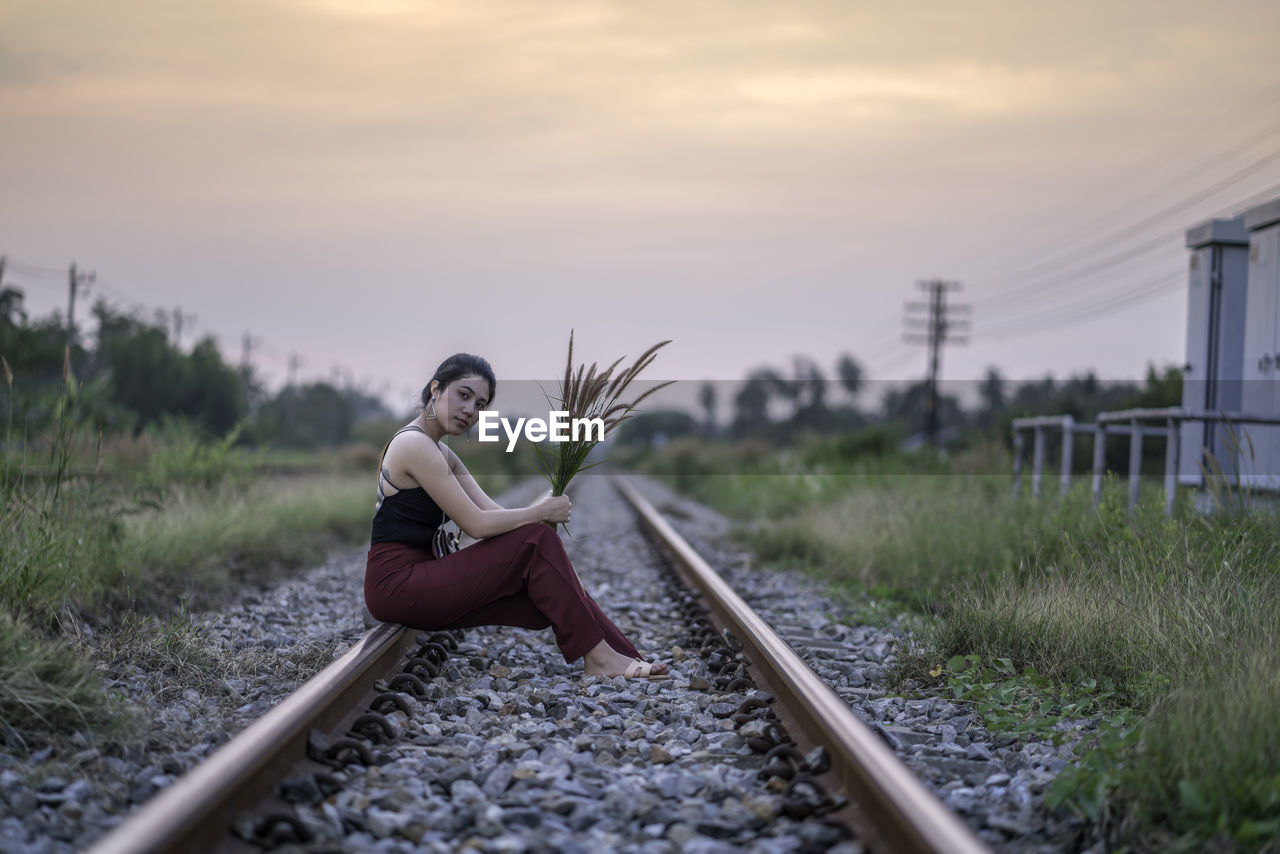 Portrait of woman holding plant while sitting on railroad track during sunset