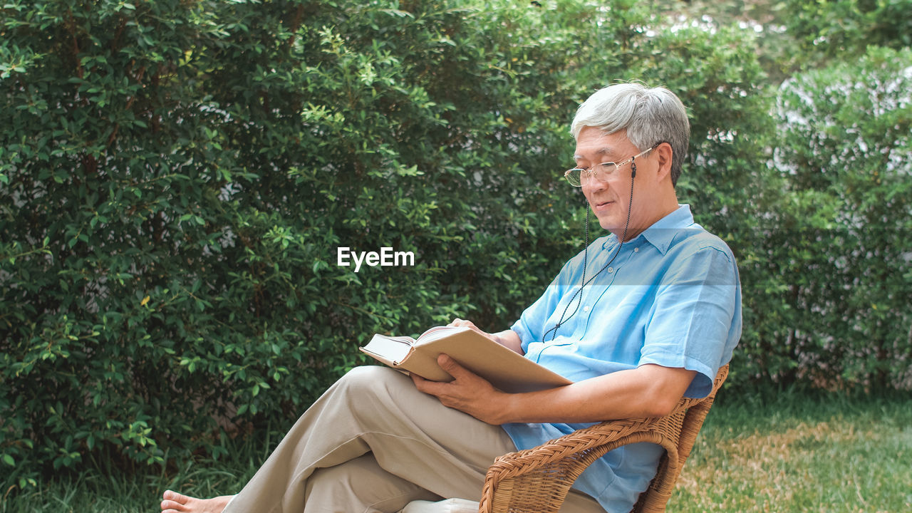 Side view of man reading book while sitting on chair against plants