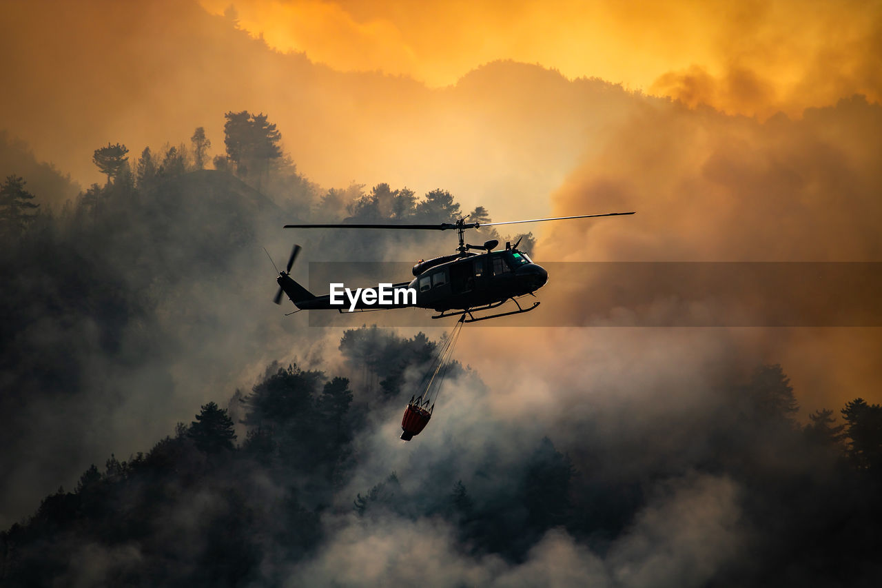 Low angle view of silhouette helicopter flying against smoke during sunset