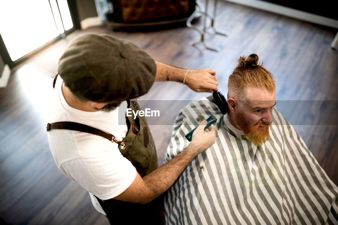 From above modern hairdresser barber cutting a adult man redhead's hair in barber chair