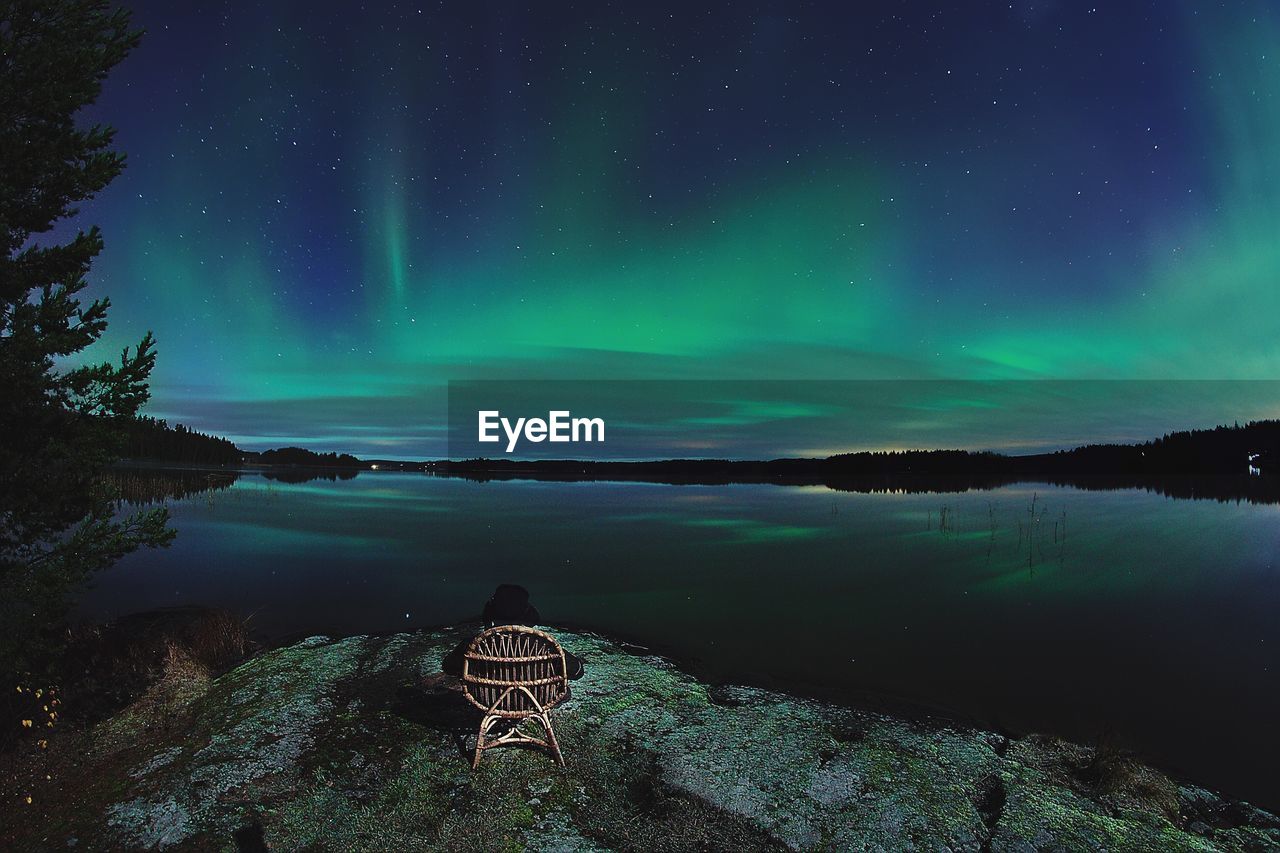 Rear view of person sitting on chair by lake against aurora polaris in sky