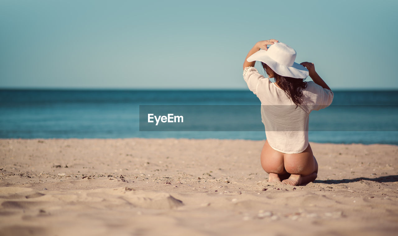 Rear view of seductive woman kneeling at beach during sunny day