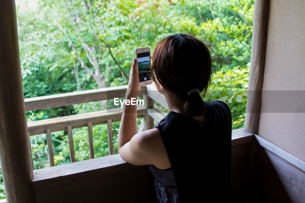 Woman photographing through mobile phone while standing by window